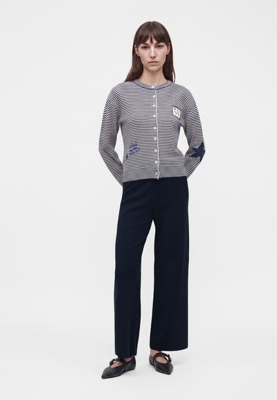 Femme portant un cardigan boutonné rayé bleu marine et blanc avec des patchs, un pantalon large bleu marine et des ballerines noires pointues avec des brides à la cheville.