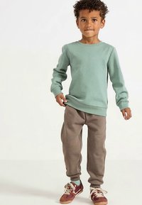 Felpa verde chiaro a maniche lunghe abbinata a pantaloni della tuta marroni. Il bambino indossa sneakers bordeaux e crema con i lacci.