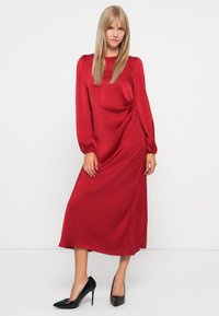 Robe longue en satin rouge à manches longues avec une taille ajustée et un détail drapé sur le côté. Portée avec des escarpins noirs à bout pointu. Texture lisse et brillante.