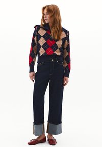 Maglione argyle blu navy e rosso con colletto alto, abbinato a jeans a gamba larga blu scuro con orli in denim e scarpe rosse.