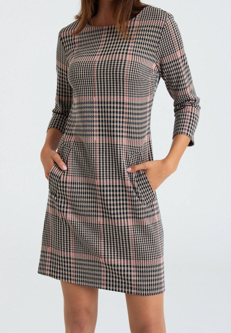 Femme portant une robe mi-longue à manches longues avec un motif pied-de-poule et à carreaux noir, blanc et rose, mains dans les poches avant.