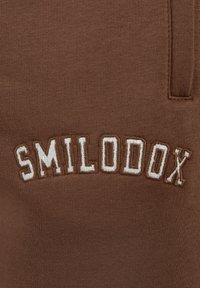 Brązowy materiał bluzy z wyszywanym napisem "SMILODOX" w kremowym kolorze. Teksturowana struktura z gładkim wykończeniem i detalem w postaci bocznej kieszeni.