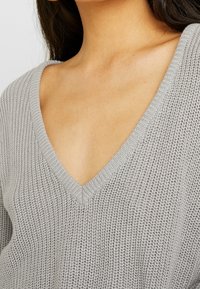 Maglione grigio in maglia con un profondo scollo a V, caratterizzato da una finitura testurizzata e bordi a costine lungo il collo e l'orlo.
