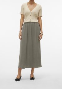 Cardigan de manga corta beige claro con tejido acanalado y botones, combinado con una falda larga y vaporosa de color verde oliva y zapatos de tacón negros con punta en forma de pico.