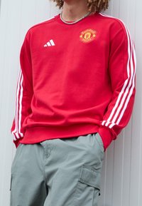 Persona che indossa una felpa rossa del Manchester United con strisce bianche sulle maniche e pantaloni cargo grigi, con le mani nelle tasche, contro un muro chiaro.