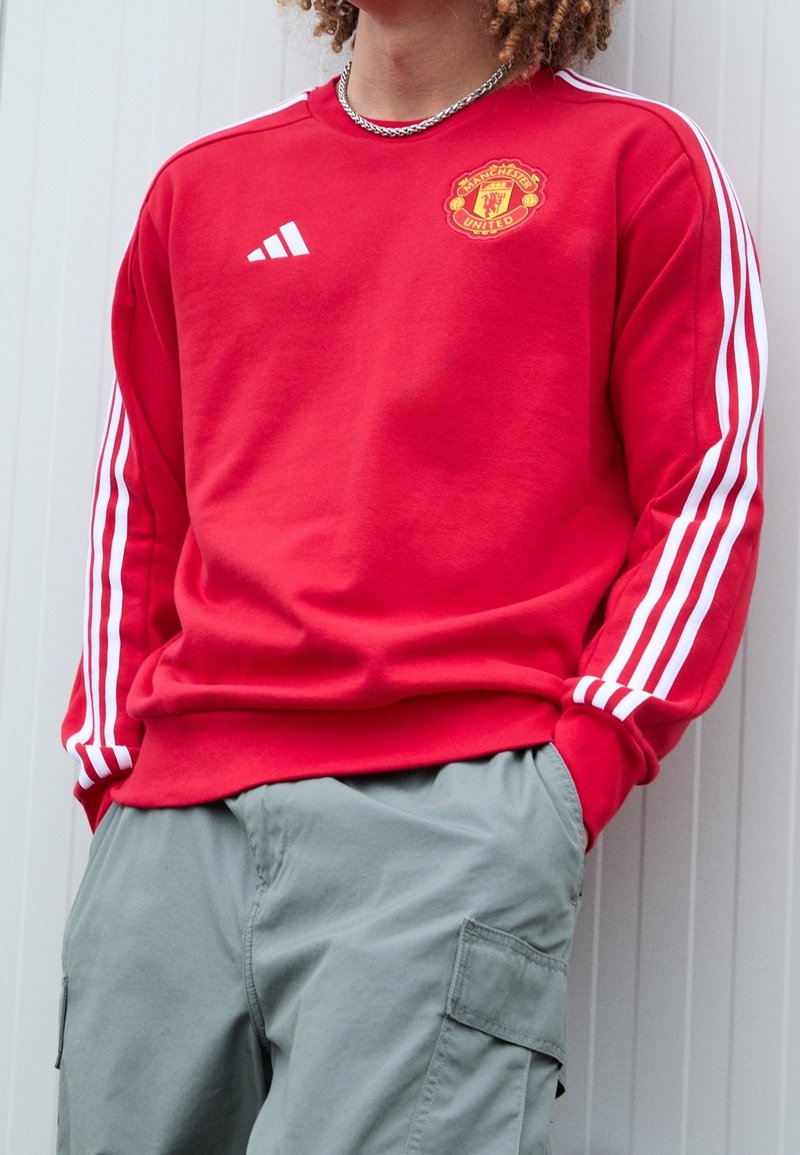 Persona che indossa una felpa rossa del Manchester United con strisce bianche sulle maniche e pantaloni cargo grigi, con le mani nelle tasche, contro un muro chiaro.