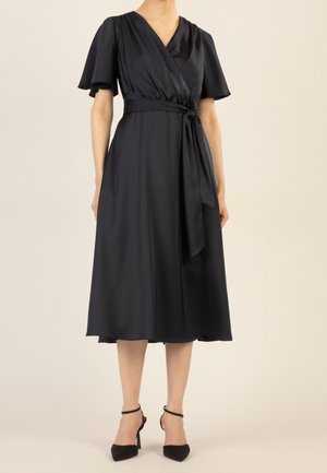 Femme portant une robe portefeuille midi noire avec des manches courtes volantées et une ceinture nouée à la taille, associée à des talons noirs à bride cheville.