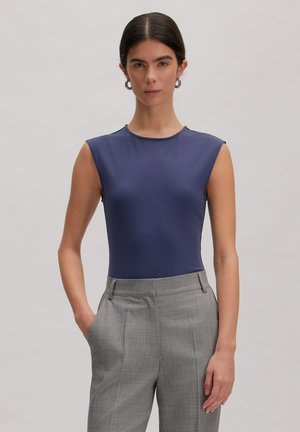 Frau mit dunklem Haar in einem marineblauen ärmellosen Oberteil und hoch taillierter grauer Hose, die mit einer Hand in der Tasche vor einem einfarbigen Hintergrund steht.