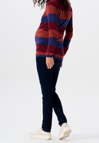 Svetr s dlouhým rukávem, pruhovaný v burgundské, oranžové a námořnické modři, doplněný tmavými skinny džíny a světlými teniskami, zobrazený z boku.