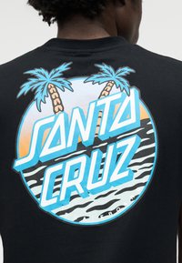 Henkilön takaosa, joka pukeutunut mustaan paitaan, jossa on värikäs pyöreä Santa Cruz -logo, palmupuut, auringonlasku ja meren aallot -kuvio.