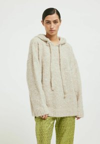Pulover bej, pufoș, cu glugă, de dimensiuni largi, cu șnururi la guler și mâneci lungi. Asortat cu pantaloni verzi, strălucitori, cu un model.