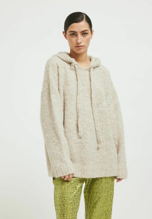 Pulover bej, pufoș, cu glugă, de dimensiuni largi, cu șnururi la guler și mâneci lungi. Asortat cu pantaloni verzi, strălucitori, cu un model.