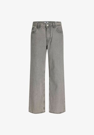 Adolescent portant une veste en jean foncée, un jean gris clair, des chaussures noires, un t-shirt beige et une casquette grise, debout et souriant devant un fond uni.