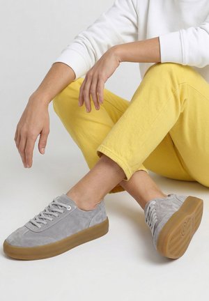 Persoon zittend met gekruiste benen, gekleed in een gele broek, wit shirt met lange mouwen en grijze suède sneakers met bruine zolen op een witte achtergrond.