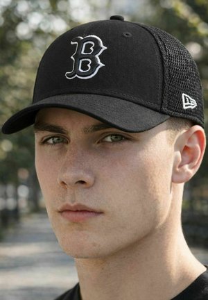 Junger Mann mit schwarzer Baseballkappe und einem markanten weißen "B"-Logo, fotografiert im Freien vor einem verschwommenen natürlichen Hintergrund.