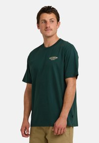 Camiseta de algodón verde oscuro con mangas cortas, que presenta un pequeño logo impreso en el área del pecho izquierdo y un corte relajado.