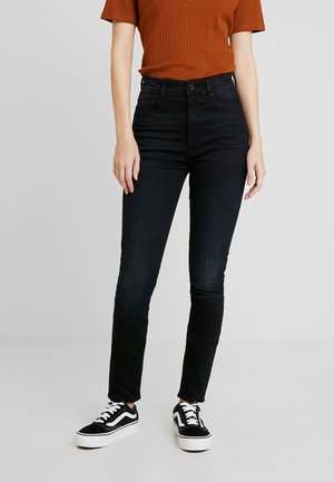 Jeans negros de cintura alta en un corte ajustado, confeccionados en denim con una textura suave, combinados con zapatillas negras y blancas.
