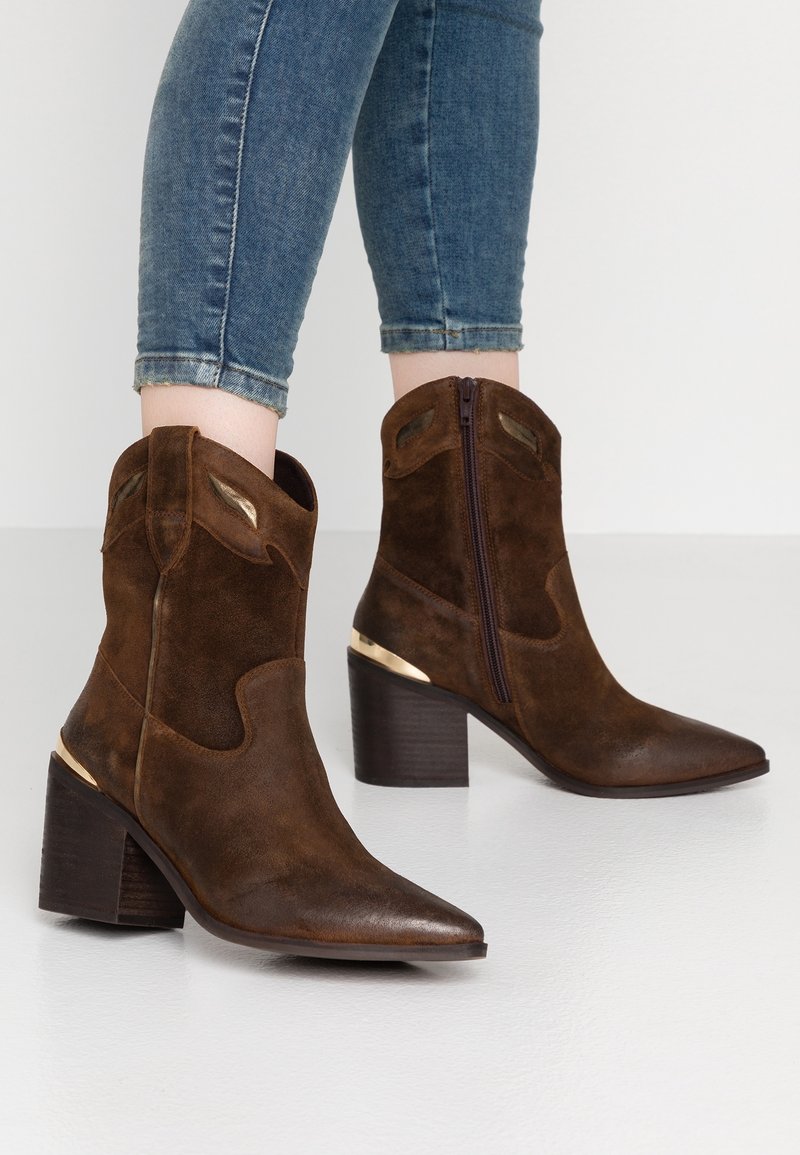 Hnědé semišové kotníkové boty s špičatou špičkou, zlatým akcentem na podpatku a bočním zipem, vystavené s modrými džínami na bílém pozadí.