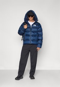 Doudoune bleu marine, avec une capuche, fermeture zippée, et un logo sur la poitrine. Associée à un pantalon noir et des baskets foncées.