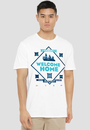 Man wearing white T-shirt with blue Hogwarts castle graphic and "Welcome Home" text, standing against plain gray background.