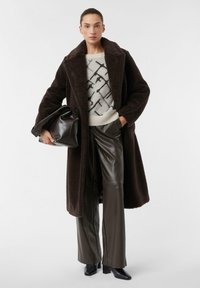 Cappotto di pelliccia sintetica marrone con collo a revers, indossato su un maglione grigio chiaro con un motivo astratto nero, abbinato a pantaloni di pelle marrone scuro e stivaletti neri con tacco.