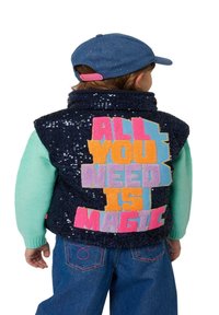 Marineblauwe pailletten vest met kleurrijke stoffen letters die "ALL YOU NEED IS MAGIC" lezen, gedragen over een mintgroen shirt met lange mouwen, gecombineerd met denim jeans.