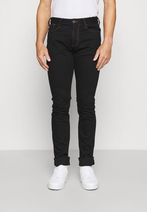 Personne portant un jean noir retroussé, des baskets blanches et une chemise blanche, debout devant un fond clair uni.