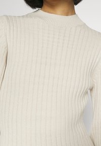 Maglione a coste in beige chiaro, caratterizzato da un design aderente, collo alto e maniche lunghe. Tessuto strutturato con una leggera lucentezza.