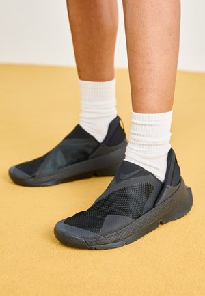 Fekete slip-on cipők, hálós és gumis részletekkel, textúrázott talppal és sima felsőrésszel, fehér bordás zoknival párosítva.