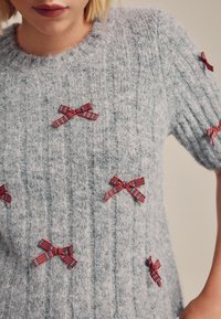 Pull en tricot gris avec texture côtelée, orné de plusieurs nœuds rouges et à carreaux cousus sur le tissu pour la décoration.