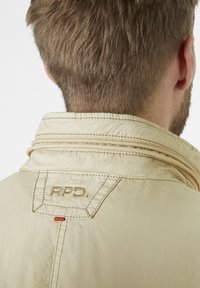 Beige jacket with a folded collar, featuring textured fabric and visible stitching. "RPD" logo appears on the collar.