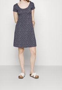 Sukienka z krótkim rękawem i okrągłym dekoltem z ciemnym tłem w wielokolorowy wzór w plamki, w zestawieniu z białymi sandałami na krzyż.