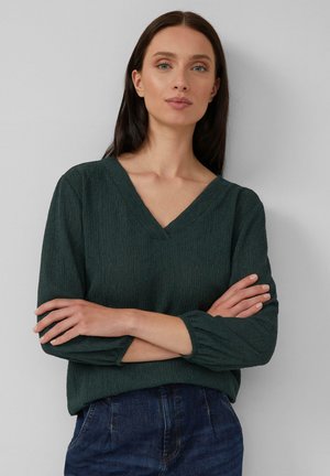 Dunkelgrünes, strukturiertes V-Ausschnitt-Oberteil mit langen Ärmeln und lockerer Passform, kombiniert mit blauen Denim-Jeans. Einfaches und lässiges Design.
