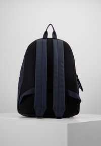 Musta ja tummansininen reppü, jossa on pehmustetut olkahihnat ja verkkoselkäpaneeli, lepää valkoisella jalustalla tasaisen harmaan taustan edessä.