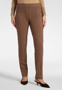 Bruine op maat gemaakte broek met een gladde stofafwerking, met een slim fit en subtiele vouw aan de voorkant. Gecombineerd met donkerbruine hakken.