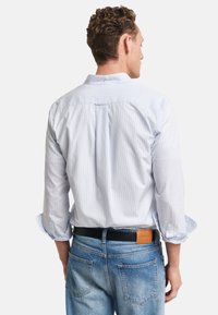 Hellblau gestreiftes Hemd mit Knopfleiste am Kragen, umgeschlagenen Manschetten und einem zentralen Rückenfalz, getragen zu blauen Jeans aus Denim mit einem Lederpatch.