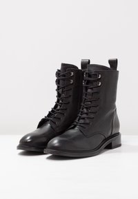 Bottines en cuir noir avec une texture lisse, lacets à l'avant, fermeture éclair sur le côté et boucle arrière. Talon bas empilé et détails de couture.