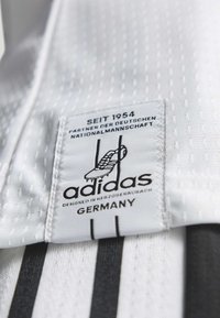 Tissu de maillot blanc avec surface texturée. L'étiquette Adidas présente un texte noir, un graphique de chaussure et des rayures verticales. Conçu en Allemagne.