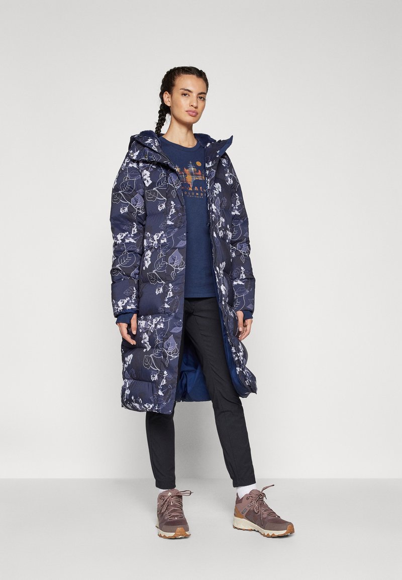 Manteau long en duvet bleu marine avec un motif floral blanc, capuche et tissu texturé ; porté sur un t-shirt graphique bleu et un pantalon noir avec des bottes de randonnée marron.