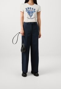 Camiseta blanca con logo triangular de flores azules y texto "GUESS CALIFORNIA", combinada con pantalones de pierna ancha de talle alto en color azul marino y un bolso negro.
