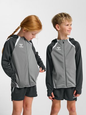 To børn iført grå og sorte sportsjakker og shorts; den ene ser ned og justerer jakken, den anden smiler og kigger mod højre.