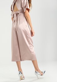 Saténový overal v jemně růžové barvě s hlubokým výstřihem na zádech, polovičními rukávy a zavazováním v pase, doplněný o kovově stříbrné vysoké podpatky.