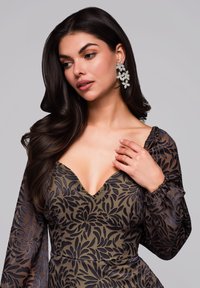 Blumengemustertes Kleid mit langen, transparenten Ärmeln in Olivgrün und Dunkelblau. Verfügt über einen tiefen V-Ausschnitt und einen strukturierten Stoff, ergänzt durch blumenförmige Ohrringe.