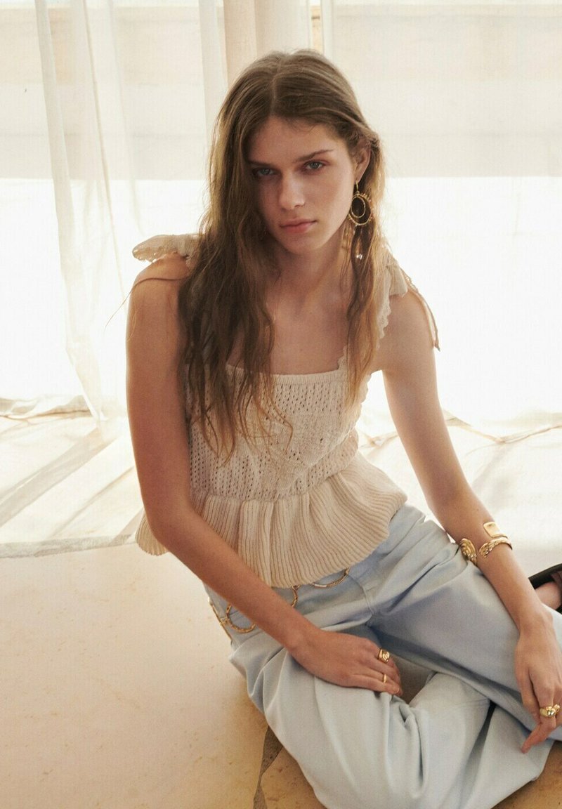 Jeune femme aux longs cheveux portant un haut beige en tricot sans manches, un pantalon bleu clair et des bijoux en or, assise dans une lumière naturelle douce.