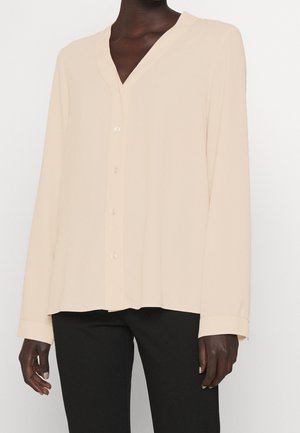 Personne portant un chemisier crème à manches longues boutonné avec un col en V et un pantalon noir, debout devant un fond uni.