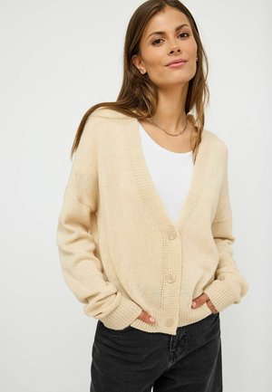 Cardigan beige lavorato a maglia con scollo a V, chiusura con bottoni e polsini a coste. Indossato sopra una T-shirt bianca con vestibilità rilassata e tessuto testurizzato.