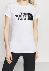 Bílý bavlněný tričko s černým nápisem "THE NORTH FACE" na přední straně, doplněné černým logem polokoule. Krátké rukávy, standardní střih.
