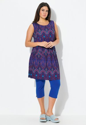 Mouwloze paarse jurk met geometrisch patroon, gecombineerd met capri-legging in het blauw. Accessoires zijn blauw instap schoenen.