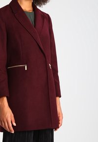 Burgundyröd ullblandad kappa med en figursydd design, notched krage och två dragkedjefickor. Slät yta, medellång, med skräddarsydda ärmar.