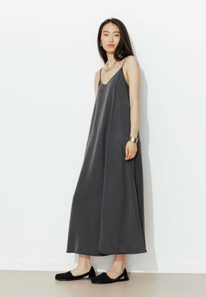 Femme portant une robe sans manches, longue jusqu'aux chevilles, gris foncé, avec des chaussures plates noires, debout contre un mur blanc uni.